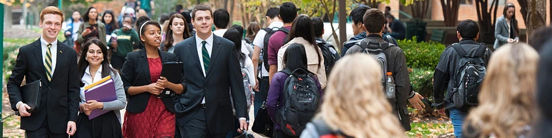 A group of students, some in formal attire and others casually dressed, walk together on a college campus path surrounded by greenery.