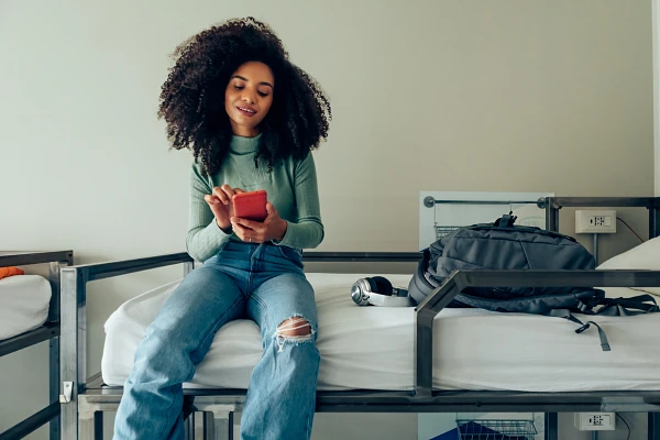 A woman with curly hair sits on a bed using her phone, with a backpack and headphones next to her.
