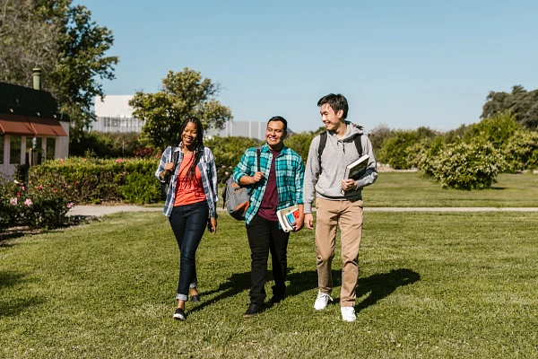 Three friends are walking and chatting in a park on a sunny day, carrying books and backpacks.